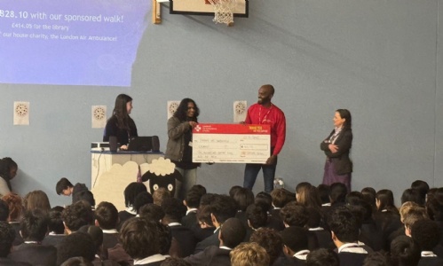 House captains handing over a large cheque to London Air Ambulance representative, in front of the Lamb students during their assembly